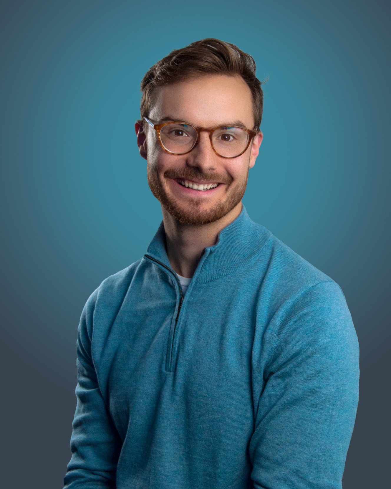 Isaac Miller smiling in glasses and a teal quarter-zip against a blue studio backdrop