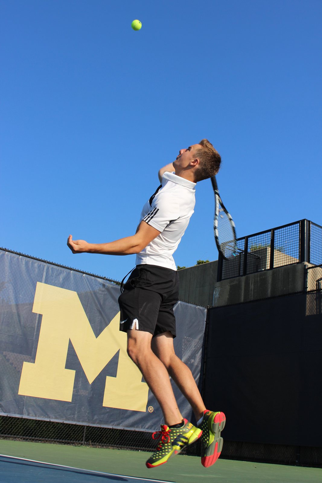 Isaac mid-serve on a tennis court with a block M banner behind